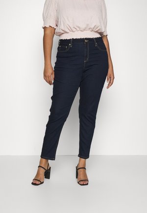 Vrouw draagt donkerblauwe high-waist slim-fit jeans, lichtroze top met pofmouwen en zwarte sandalen met hak, staand tegen een effen witte achtergrond.