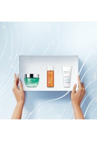 Trzy produkty do pielęgnacji skóry Biotherm na białej tacy: zielony słoik, pomarańczowa butelka i biała tubka, na jasnoniebieskim tle.