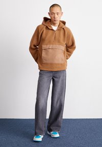 Pulôver de fleece castanho com capuz, fecho de zíper frontal até metade e um grande bolso bege. Combinado com jeans de perna larga cinzentos e sapatos com detalhes azuis.