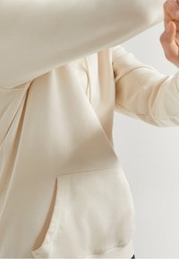 Krémově zbarvená mikina s kapucí z měkké látky, vybavená klokáním kapsou a žebrovanými lemy na rukávech. Design má volný střih a hladkou texturu.