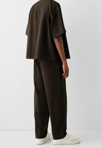 Hnědý oversized top s krátkým rukávem, rovný lem a uvolněný střih, ve spojení s volnými kalhotami ve stejném odstínu a krémovými teniskami.