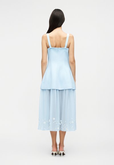 Femeie cu păr lung și închis la culoare, purtând o rochie cu volane fără mâneci, de culoare albastru deschis, cu broderii florale lângă margine și pantofi cu toc albi, stând cu spatele către cameră.