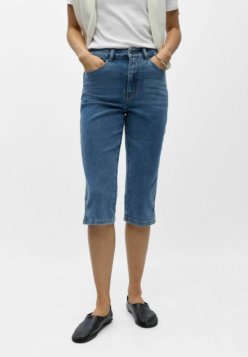 Personne portant un short en jean bleu mi-long, une chemise blanche, un gilet sans manches crème, des chaussures noires sans lacets, debout les mains dans les poches.