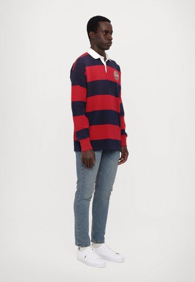 Tricou polo cu dungi roșii și bleumarin, cu mânecă lungă și guler alb, purtat cu blugi de culoare albastru deschis și adidași albi. Prezintă un logo pe piept.