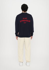 Sweatshirt azul-marinho com texto em vermelho "MILANO GYMNASIUM EA7" nas costas. Punhos e bainha canelados. Combinado com calças creme e sapatilhas brancas.