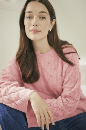 Hellrosa gestrickter Pullover mit Rautenmuster und gerippten Bündchen, kombiniert mit blauen Jeans. Das Model posiert sitzend mit einem entspannten Ausdruck.