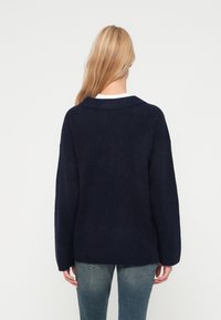 Sötétkék, oversized pulóver bordázott gallérral és mandzsettával, puha tapintású anyagból készült, fehér ing felett viselve és farmernadrággal párosítva.