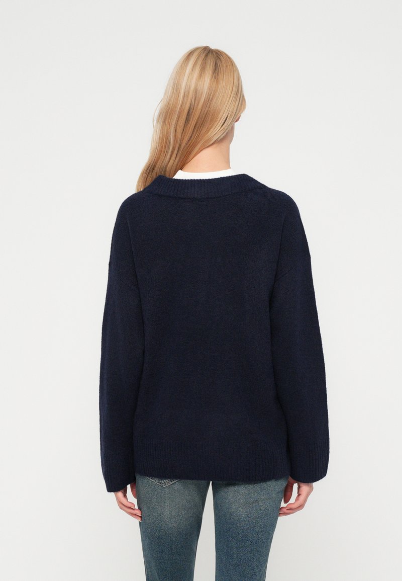 Sötétkék, oversized pulóver bordázott gallérral és mandzsettával, puha tapintású anyagból készült, fehér ing felett viselve és farmernadrággal párosítva.