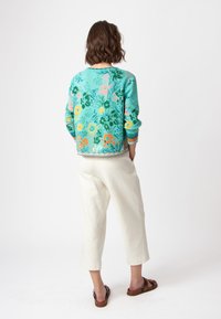 Suéter con estampado floral en turquesa, que presenta tonos de verde, rosa y naranja. Combinado con pantalones de corte relajado de color claro y sandalias.