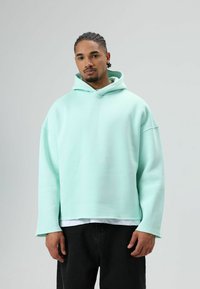 Sweat à capuche oversize vert menthe en tissu doux avec poche kangourou, épaules tombantes et ourlet brut, associé à un jean noir.
