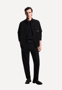 Giovane uomo in piedi, vestito con un completo totalmente nero composto da camicia con bottoni, maglia a maniche lunghe, pantaloni, cintura nera e mocassini neri su sfondo bianco.