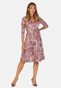 Pitkähihainen, paisley-kuvioinen mekko vaaleanpunaisena ja ruskeana, jossa on kietaisumalli ja kireä vyötärö, täydentäen ruskeat korkokengät.