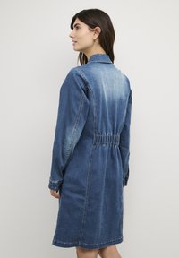 Denim kleita ar apkakles izgriezumu, piegulošu vidukli, garām piedurknēm un vidēja garuma dizainu ar izbalējušu zilo tonu.