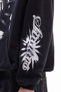 Μαύρο φούτερ με κουκούλα πο υ διαθέτει λευκό γραφικό σχέδιο στο μανίκι και μπροστά. Το υλικό φαίνεται απαλό και σαν φλις, με άνετη εφαρμογή.