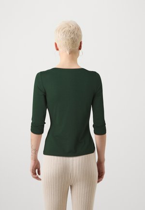 Personne aux cheveux blonds courts portant un haut vert foncé à manches 3/4 et un pantalon clair côtelé, debout face à un fond blanc uni.