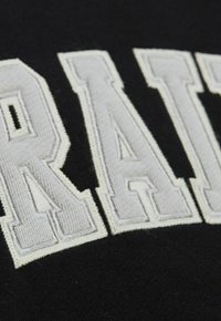 Schwarzer Stoff mit großen, weißen bestickten Buchstaben. Die Textur ist glatt und hat einen leichten Glanz auf dem Faden.