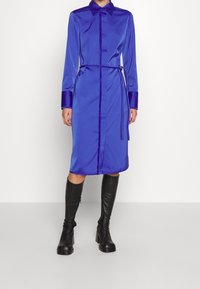 Dlouhé rukávy, šaty ke kolenům v jasně modrém saténu, s košilovým límečkem, páskem v pase a kontrastními tmavě modrými manžetami.