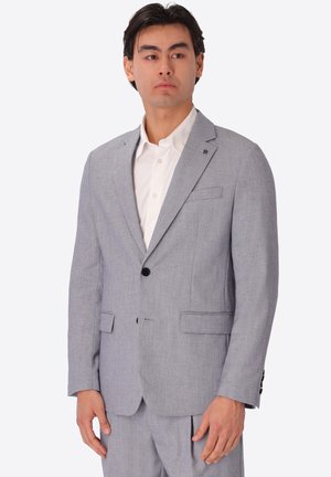Uomo che indossa giacca da completo grigio chiaro e pantaloni abbinati con una camicia bianca, in piedi su uno sfondo semplice.