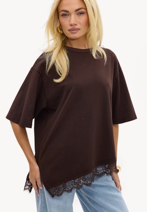 Femme blonde portant un T-shirt marron foncé oversized avec un bord en dentelle à l'ourlet, un jean bleu clair, des boucles d'oreilles créoles dorées et un bracelet en or.
