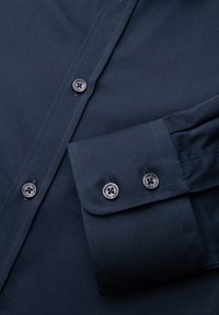 Chemise bleu foncé à manches longues montrant les boutons avant et un poignet boutonné avec deux boutons visibles sur un tissu lisse.