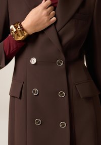 Mano con braccialetto spesso dorato che sistema un blazer doppiopetto marrone scuro con bottoni con bordo argento e tasche con pattina.