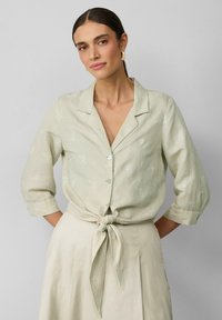Lichtgroen linnen shirt met witte bloemen borduursels, knoopsluiting en driekwart mouwen die op de taille zijn gestrikt. Glad textuur, casual ontwerp.