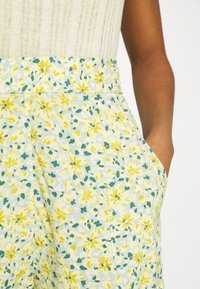 Gros plan d'une personne portant un pantalon fleur à taille haute, de couleur claire avec des fleurs jaunes et vertes, associé à un haut sans manches en tricot beige.