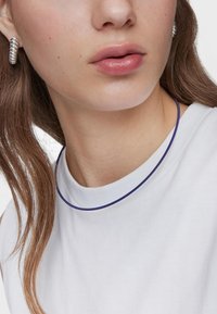 Orecchini a cerchio argento e una maglietta bianca con un dettaglio color viola sul colletto. Texture liscia, vista ravvicinata che mette in risalto i tratti del viso e i capelli.