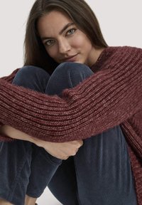 Junge Frau mit langen braunen Haaren umarmt die Knie, trägt einen bordeauxroten grob gestrickten Pullover und marineblaue Cordhosen vor einem schlichten Hintergrund.