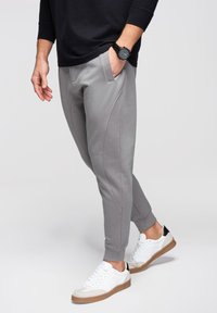 Mężczyzna ubrany w szare spodnie dresowe, białe trampki z brązowymi podeszwami, czarną koszulę z długim rękawem oraz czarny zegarek na nadgarstku, stojący z jedną ręką w kieszeni.