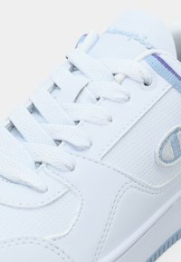 Λευκό αθλητικό παπούτσι με κορδόνια, υφασμάτινες λεπτομέρειες με υφή, μπλε και μοβ περιθώριο, και κεντημένο λογότυπο στο πλάι και στη γλώσσα.