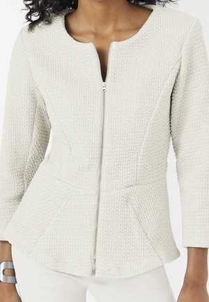 Kobieta ubrana w teksturowaną, kremowobiałą kurtkę z przednim zamkiem błyskawicznym i rękawami do łokcia, w połączeniu z białymi spodniami i srebrną bransoletką.