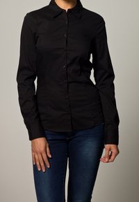 Camicia nera a maniche lunghe con bottoni, caratterizzata da un colletto, design aderente e tessuto liscio e anti-piega. Abbinata a jeans blu.