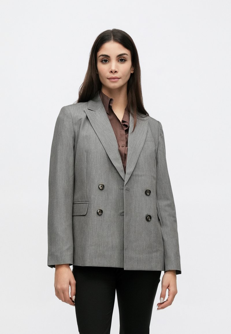 Blazer gris croisé avec un revers à encoche, tissu texturé, boutons noirs, et deux poches avant, porté sur une chemise marron.