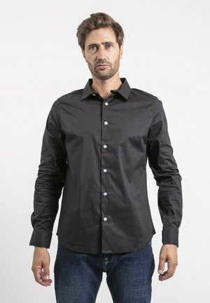 Homme aux cheveux châtain clair portant une chemise noire boutonnée et un jean bleu, debout devant un fond blanc uni, faisant face à l'avant.