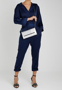 Jumpsuit in satin blu navy con design avvolgente, cintura abbinata e polsini arricciati. Abbinato a una clutch argentata e tacchi neri con dettagli floreali.