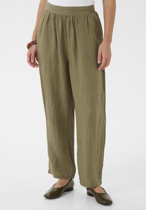 Luźne lniane spodnie w oliwkowym kolorze na osobie noszącej brązowe baleriny i biały top, stojącej na jednolitym tle.