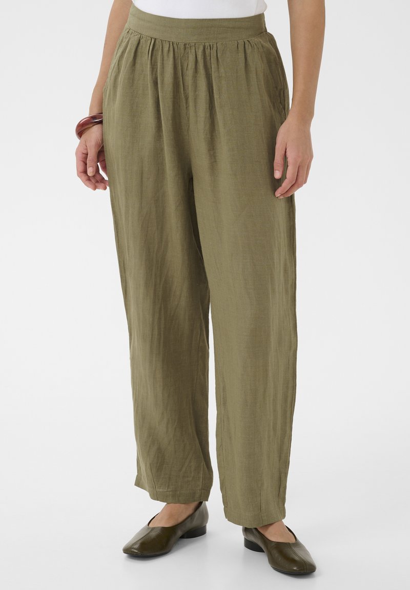 Pantalon ample en lin vert olive porté par une personne chaussant des ballerines marron et un haut blanc, debout devant un fond uni.