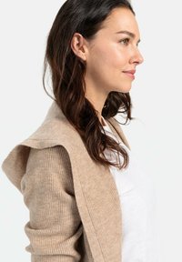 Beige Rippstrick-Cardigan mit Kapuze, weicher Textur und offener Vorderseite. Getragen über einem weißen T-Shirt, präsentiert vor einem neutralen Hintergrund.