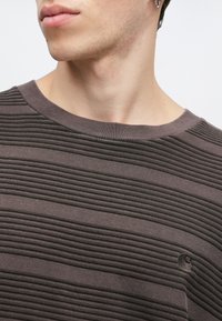 Suéter texturizado de color marrón oscuro con estrías horizontales, cuello redondo y un pequeño logo en la esquina inferior. Detalle de tela suave visible.