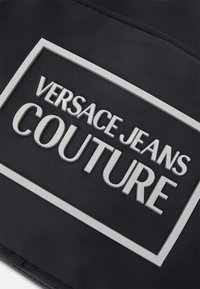 Svart tyg med upphöjd vit text "VERSACE JEANS COUTURE" inuti en rektangulär ram, vilket betonar en slät textur och modern design.
