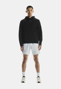 Zwarte hoodie met een frontzak, gecombineerd met lichtgrijze shorts. Model draagt witte sportschoenen met zwarte accenten.