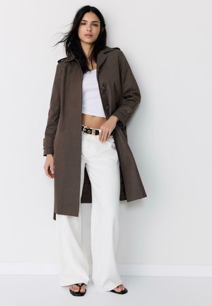 Donna con un trench marrone sopra un crop top bianco, pantaloni bianchi a gamba larga, cintura nera con fibbia dorata e sandali neri, in piedi contro un muro bianco.