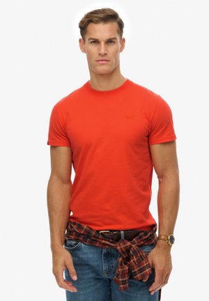 Jeune homme portant un t-shirt rouge ajusté, un jean bleu et une chemise à carreaux nouée autour de la taille, debout devant un fond blanc.