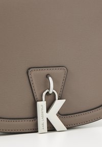 Brązowa torebka o teksturowanej powierzchni z czarnym przeszyciem oraz srebrnym zawieszką z literą 'K' przymocowaną na pierścionku. Minimalistyczny design, z geometrycznym kształtem.