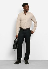 Mann, der ein beiges Hemd mit Knöpfen und schwarze Hose trägt, hält eine schwarze Aktentasche und steht vor einer schlichten weißen Wand.