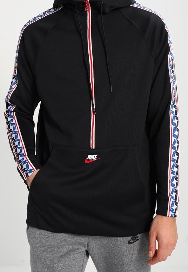 Svart Nike-huvtröja med dragkedja framtill, kontrasterande röd dragkedja och grafiskt mönster längs ärmarna. Inkluderar en framficka.