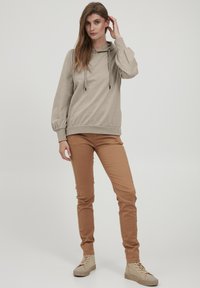 Bež jopič s kapuco in balonastimi rokavi, v kombinaciji s tan slim-fit hlačami. Model nosi bež visokotopovne športne copate. Minimalna ozadje.