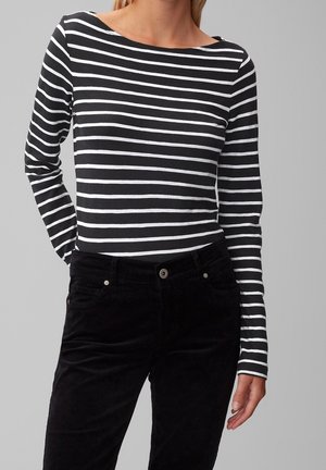 Kobieta ubrana w czarno-biały, poziomo paskowany top z długim rękawem i czarne, wysokiej talii spodnie, stojąca na jednolitym szarym tle.