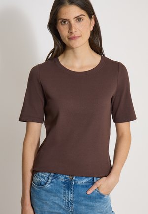 Femme aux longs cheveux foncés portant un haut marron à manches courtes et un jean bleu, la main droite dans la poche, sur un fond uni.